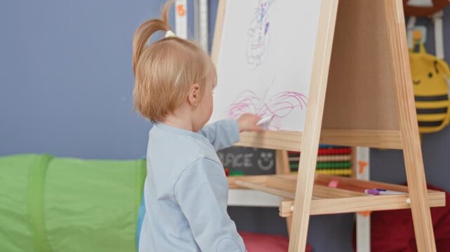 Toddler girl scribbling with marker on wooden easel in preschool, hand on whiteboard and pink scribbles visible; concentration quiet observation.
