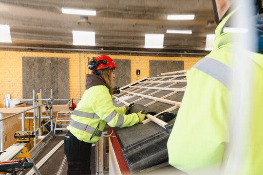 Female vocational trainee positioning concrete tile onto slanted wooden roofing battens in training workshop at secondar