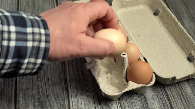 A hand in a checkered shirt reaches into an egg carton, placing one egg while several others remain visible in the carton on a wooden table in a brightly lit kitchen environment