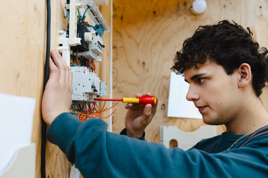 Focused vocational male student using screwdriver on complex training circuit board breaker assembly in workshop at scho