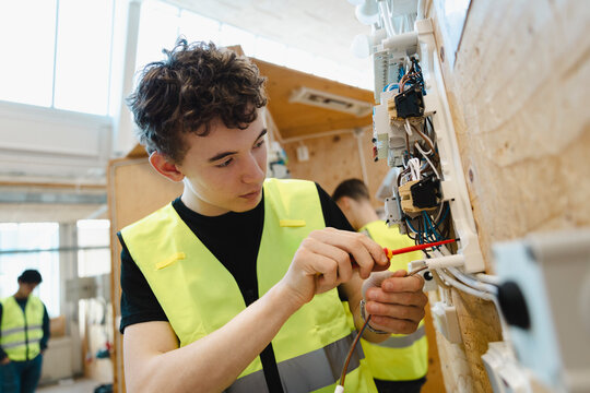 Young male student tightening components with screwdriver on wall mounted electrical breaker panel in training workshop