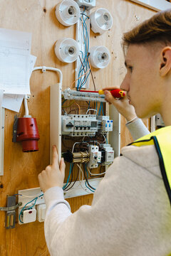 Rear view of young male student using screwdriver while installing wiring inside wall mounted electrical breaker panel a