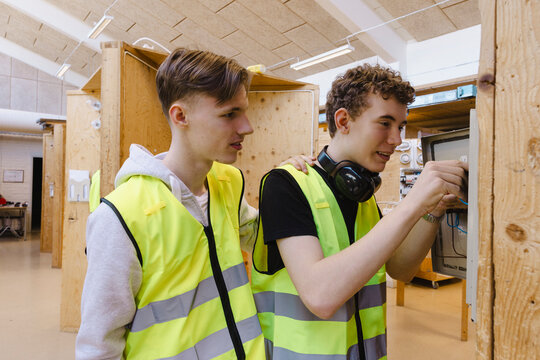 Two vocational students wearing reflective vest working together while connecting wiring inside open electrical control