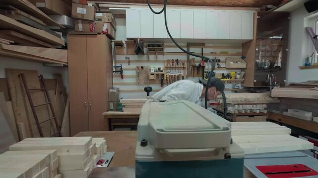 Focused male carpenter in overalls stacking light wooden pine blanks on a workbench behind a heavy thickness planer machine during a smooth camera pan in an authentic local carpentry workshop