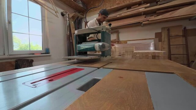Focused male fabricator examining and organizing light pine timber pieces behind a blurred thickness planer on a sturdy workbench during a smooth right to left wide angle tracking slider shot