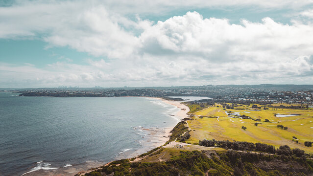 Aerial view of Long Reef Headland featuring a coastal golf course, sandy beach, and ocean waves under a cloudy sky in Collaroy, New South Wales, Australia.