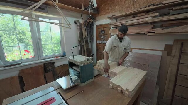 Wide angle view of a focused male carpenter in white overalls sorting and inspecting stacked wooden pine blanks on a large workbench near a thickness planer in a bright authentic carpentry workshop