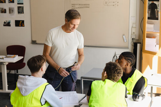 Male professor holding short piece of wire while guiding students on coursework in classroom at vocational secondary sch