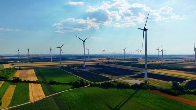 Wind turbines scattered across rural farmland from above. Aerial view of multiple turbines and dirt path crossing brown and green agricultural fields