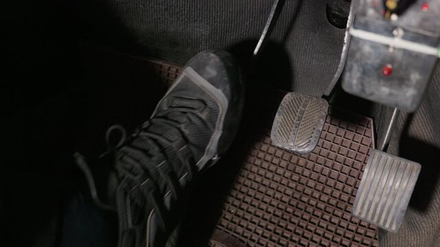 A person foot in a dark shoe presses the clutch pedal inside a manual vehicle. The floor features textured rubber pads on the brake and gas controls. Dim lighting fills the cabin.
