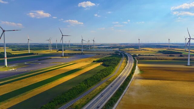 Wind turbine beside road through green farmland aerial. Drone shot of turbine and rural road crossing diagonal fields, scattered clouds above