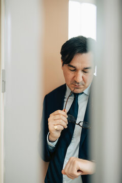 Businessman holding eyeglasses while checking time on watch in office