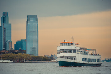 Fototapeta premium Passenger ferry on Hudson River beside Jersey City skyline. White three level vessel with people on decks, Jersey City waterfront and glass towers under cloud cover.