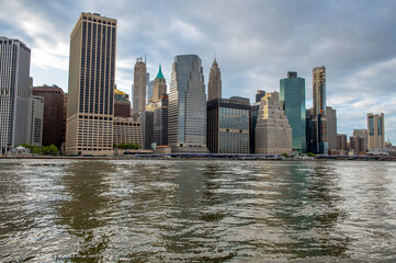 Fototapeta premium Skyscrapers of downtown Manhattan rising above New York water. Corporate office towers along financial district waterfront under cloudy sky.