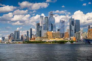 Fototapeta premium Wide skyline view of Hudson Yards towers. Cluster of skyscrapers, construction crane, waterfront buildings, cumulus clouds overhead.