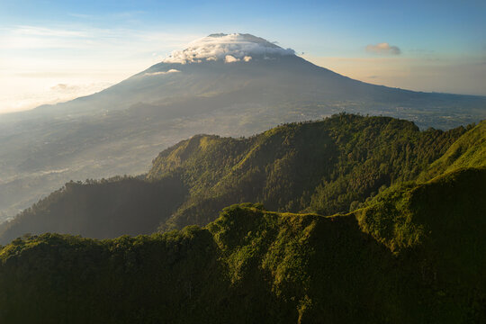 Aerial view of Telomoyo mountain with lush green slopes and a cloud-capped peak under soft golden hour light Ngablak, Central Java, Indonesia.