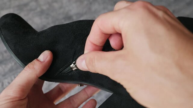 A person carefully holds and inspects a black suede heeled ankle boot in their hands. The shiny metal side zipper contrasts sharply against the dark textured fabric of the shoe.