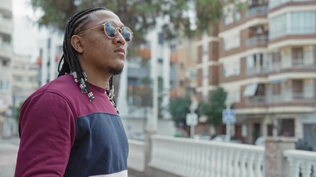 Man adjusts sunglasses with hands and looks upward on street, braided hair with beads visible; quiet contemplation.