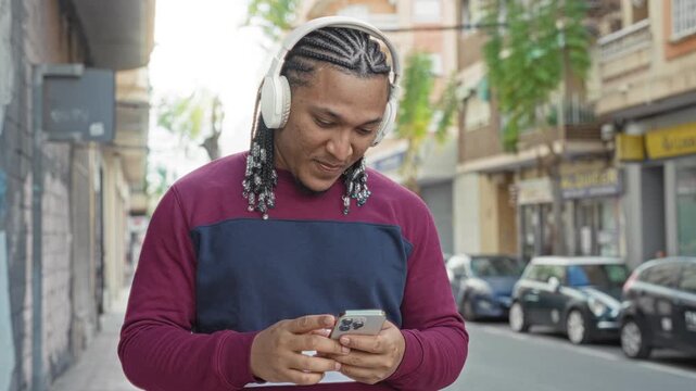 Young hispanic man wearing white headphones and a burgundy sweater tapping a smartphone with both hands while smiling on a city street lined with parked cars and shops; contentment.