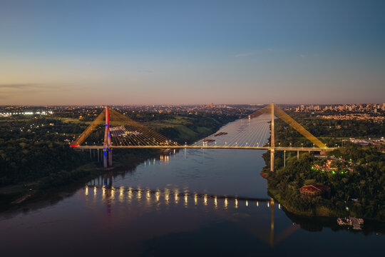 Aerial view of the Integration Bridge illuminated at dusk, spanning the Parana River with lush green banks and city lights in the background in Foz do Iguacu, State of Parana, Brazil.