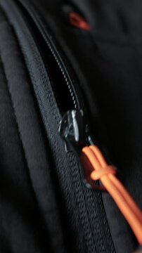 A close-up view of a black zipper slider with an orange cord puller being fastened on a dark synthetic jacket. Concept of securing gear, outdoor preparation, and tactile functionality.