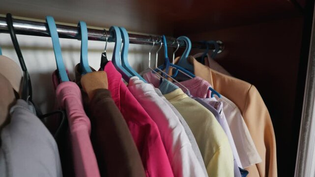 Assorted colorful garments and a peach blazer hang neatly on hangers inside a dark wooden wardrobe. Concept of daily morning routine, personal home organization, and fashion choices.