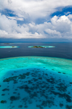 Aerial view of Dhigurah Island, Maldives, with turquoise shallows, deep navy ocean, coral islets, dark reef patches, cumulus clouds, and calm midday tropical seas.