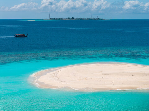 Aerial view shows Dhigurah sandbar with shallow turquoise water, deep indigo lagoon, and distant island with towers, a dhoni drifting left, under bright midday light.
