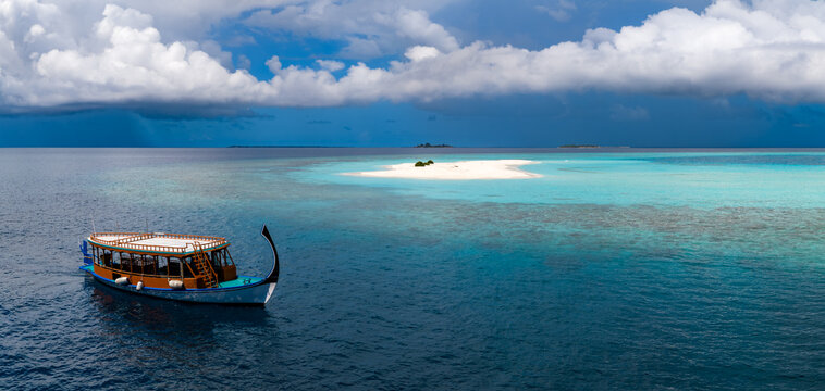 Aerial view shows a Maldivian dhoni drifting toward a tiny sandbank near Dhigurah Island, Maldives, over cobalt water and turquoise shallows under towering cumulus clouds.