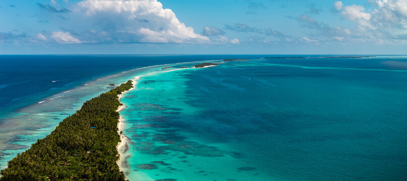 Aerial view of Dhigurah Island in the Maldives with sandspit, palm forest, white beaches, reef lagoons, and coral patches in midday light under scattered cumulus clouds.