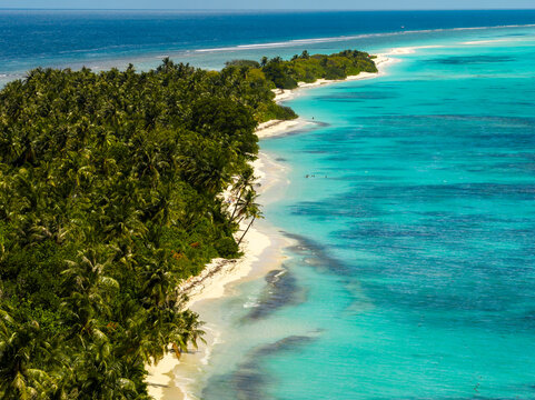 Aerial view shows Dhigurah Island, Maldives, with a long sandspit, coconut palms, turquoise shallows, a white sand beach, reef lagoon, wave lines, and coral patches at noon.