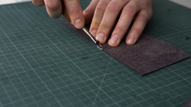 Leather craftsman beveling a leather strap with an edge creaser tool on a green cutting mat in a workshop