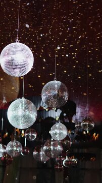 Vertical video. Mirrored disco balls hanging above a retro party with reflections and lights.