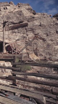 Workers are at an old mining site in a rocky area. Wooden fences surround the scene. Machinery is visible alongside weathered rock formations. The sky is clear and bright.