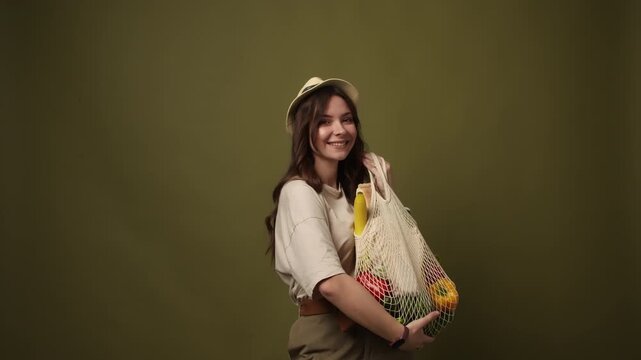 Young woman holding reusable mesh grocery bag with fresh vegetables and baguette on a green background