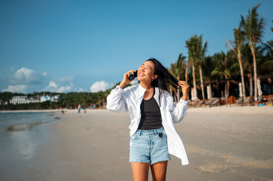 Happy young woman talking on her smartphone at a tropical beach, enjoying vacation, remote work
