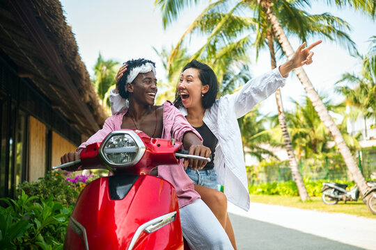Happy interracial couple riding a red vintage scooter together on a tropical island road
