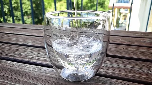 Clear drinking water pouring into glass in slow motion on wooden table. Close-up of transparent water filling a glass on balcony or terrace during sunny summer day. Hydration, healthy lifestyle