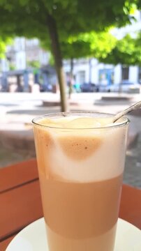 Iced latte with milk foam stirred in transparent glass at outdoor cafe. Summer lunch on restaurant terrace in a small European town square. Coffee culture, refreshment, travel, coffee shop