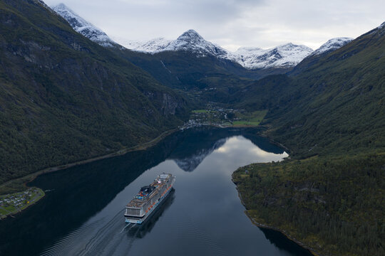 Aerial view of a large cruise ship sailing through the calm waters of Geirangerfjord, surrounded by steep mountains with snow-capped peaks and lush green slopes, Geiranger, Norway.