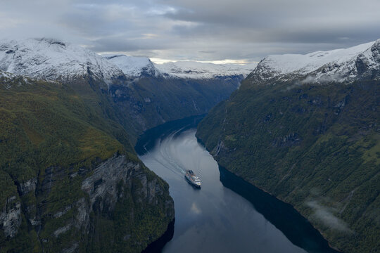 Aerial view of Geirangerfjord with a cruise ship sailing through deep blue water, flanked by steep, snow-capped mountains and green slopes in Stranda Municipality, More og Romsdal, Norway.