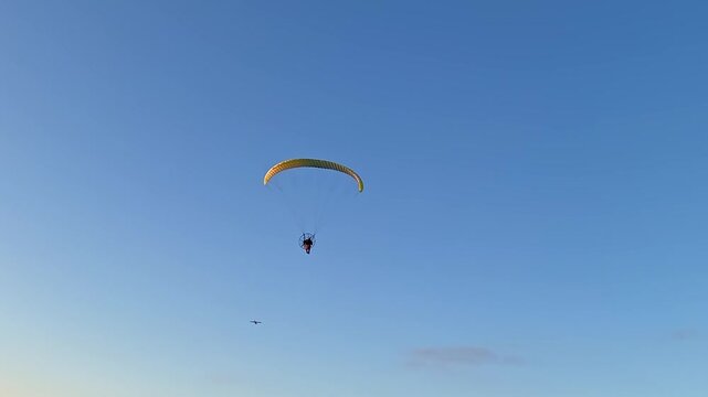 A powered paraglider pilot with a yellow canopy flies over the beach against an orange sunset.
