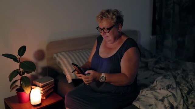 Overweight Smiling Senior Female Browsing Smartphone In Dark Bedroom Under Lamp Light