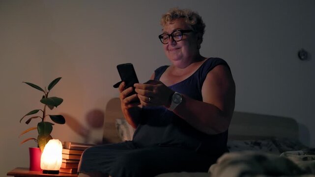 Overweight Joyful Senior Lady Holding Smartphone In Bed During Night Under Lamp Light