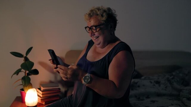 Overweight Smiling Senior Woman Texting On Smartphone In Dark Bedroom At Night By Lamp