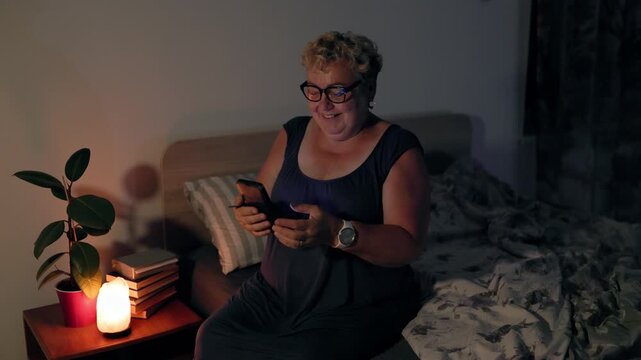 Overweight Amused Mature Woman Laughing At Smartphone Screen In Bed At Night With Lamp