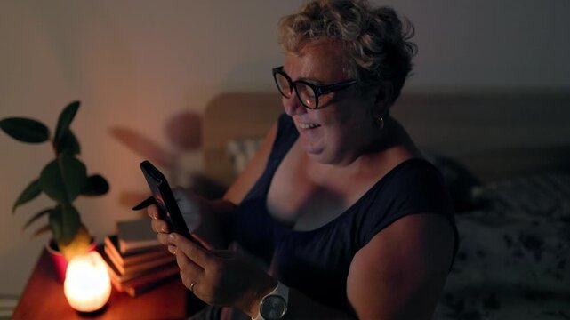 Overweight Cheerful Mature Female Browsing Social Media In Bed With Smartphone At Night