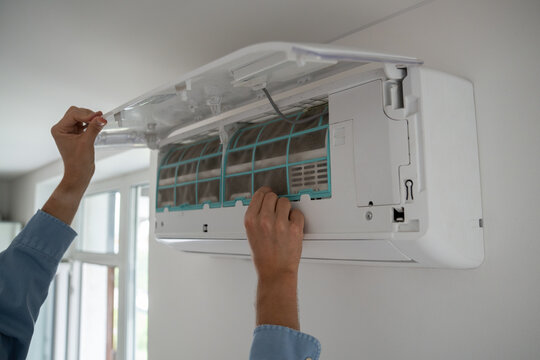 Man's hands open wall-mounted air conditioner to remove dusty filter, cooling system maintenance, male home owner cleaning indoor air conditioner unit, regular home appliance repair and service.