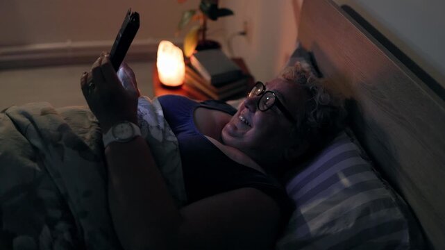 Overweight Happy Mature Woman Using Smartphone Lying In Bed At Night Under Lamp Light