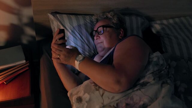 Overweight Mature Woman With Glasses Browsing Smartphone Lying In Bed At Night